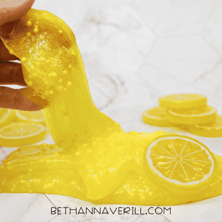 Bright yellow lemonade slime stretched upward with foam beads mixed throughout, lemon slice props nearby on a white background.