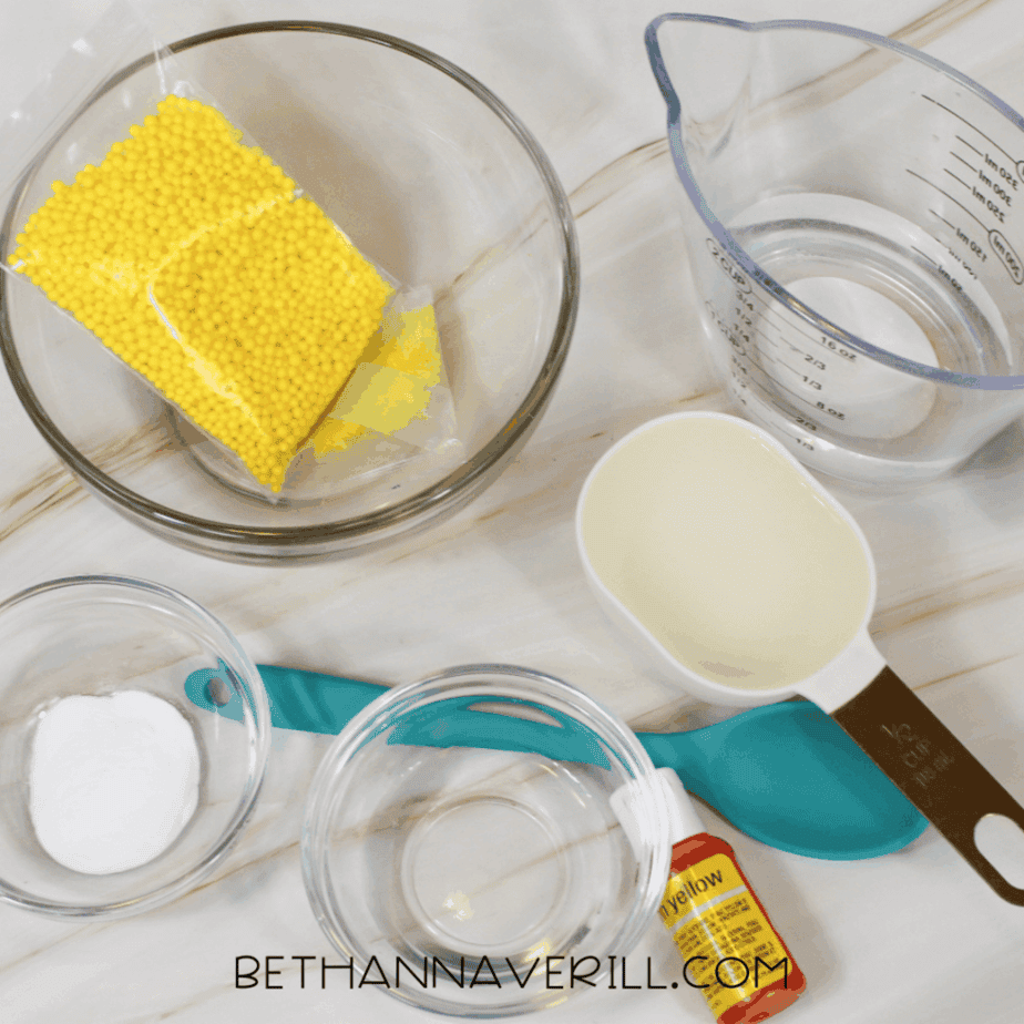Ingredients for lemonade slime arranged on a white surface, including yellow foam beads, water, glue, borax powder, measuring cups, and yellow food coloring.