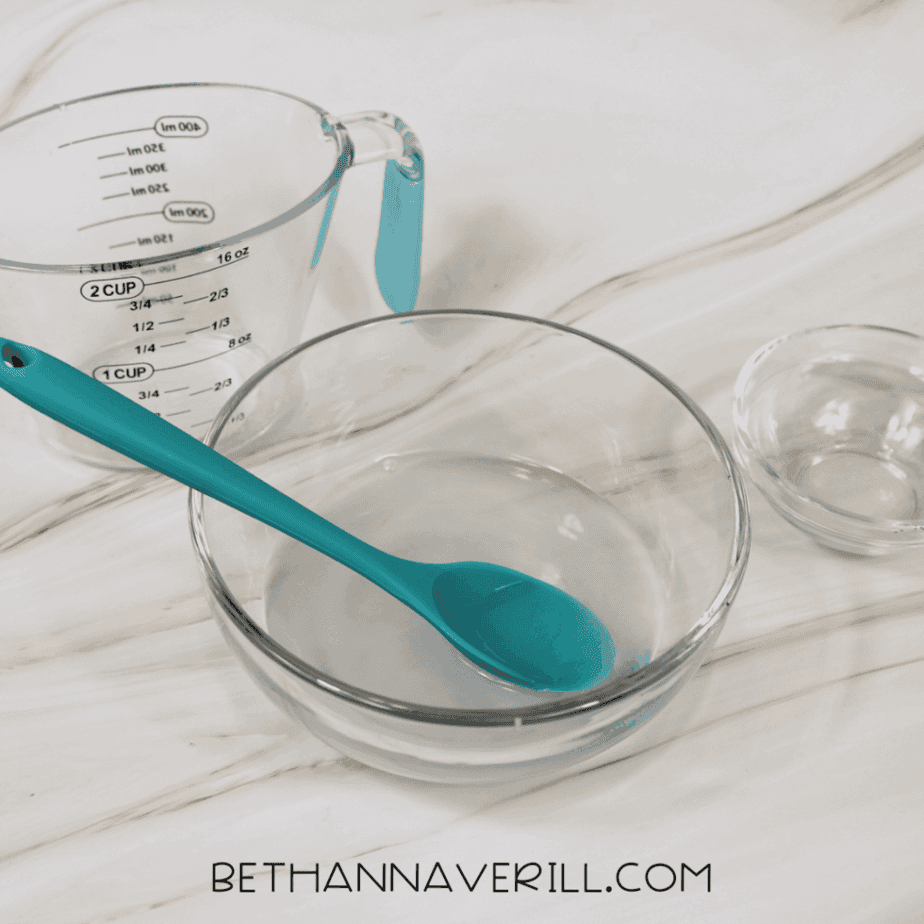 Clear bowl filled with water and a spoon resting inside, preparing liquid for lemonade slime.