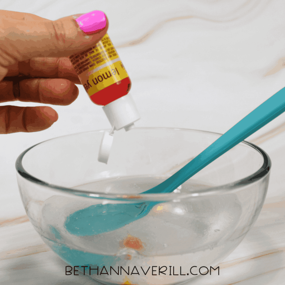 Yellow food coloring being squeezed into a bowl of clear liquid for lemonade slime.