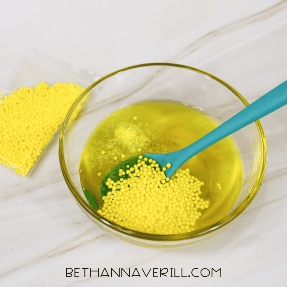 Yellow foam beads added to a bowl of yellow-tinted slime mixture and stirred with a spoon.