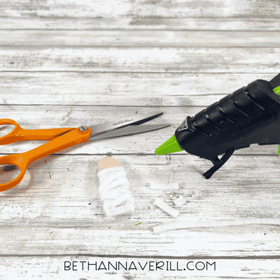 Craft stick figure wrapped in white felt strips positioned next to scissors and a hot glue gun on a white wood background.