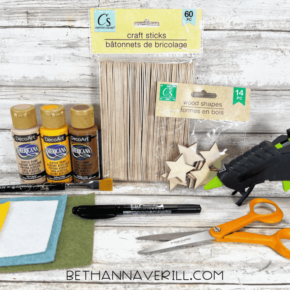 Craft supplies arranged on a white wood surface including jumbo craft sticks, wooden star shapes, acrylic paint bottles, felt sheets, pipe cleaners, scissors, a black marker, and a hot glue gun.