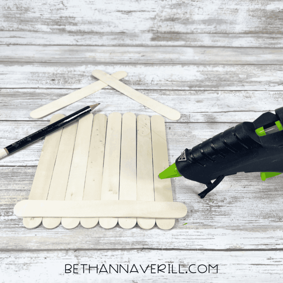 Jumbo craft sticks arranged vertically into a rectangle shape with a pencil and hot glue gun nearby on a white wood background.