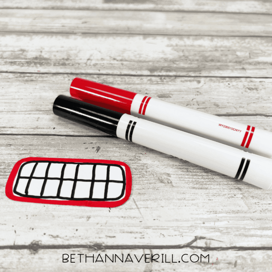 Two markers placed next to a small white cardstock piece outlined in red and black on a white wood surface.