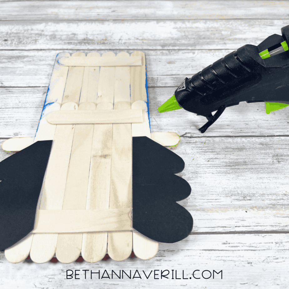 Popsicle stick base assembled with black cardstock shapes attached on both sides, with a hot glue gun on a white wood background.