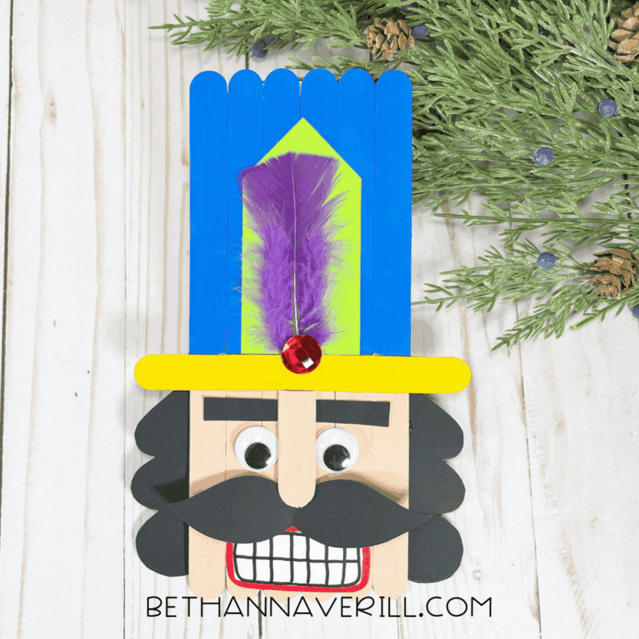 Finished popsicle stick nutcracker displayed upright on a white wood surface with green greenery in the background.