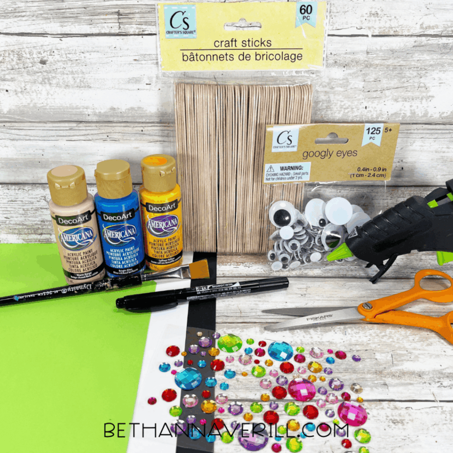 Craft supplies arranged on a white wood surface, including craft sticks, acrylic paint bottles, googly eyes, scissors, a glue gun, markers, cardstock, and colorful plastic gems.