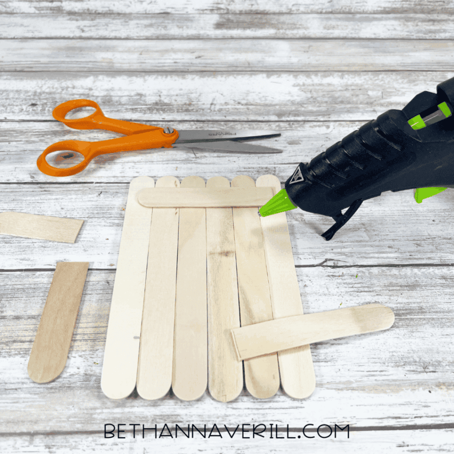 Popsicle sticks glued into a rectangle base with a horizontal stick across the top edge, with a hot glue gun nearby on a white wood background.