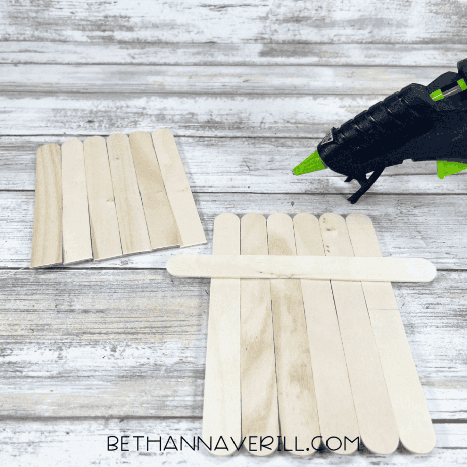 Close-up of a partially assembled craft stick rectangle with additional craft sticks placed nearby and a glue gun positioned to the right.