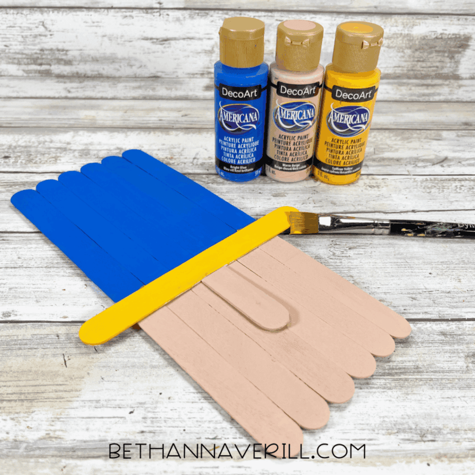Painted craft stick pieces in blue, yellow, and flesh tones drying on a white wood surface, with paint bottles and a paintbrush nearby.