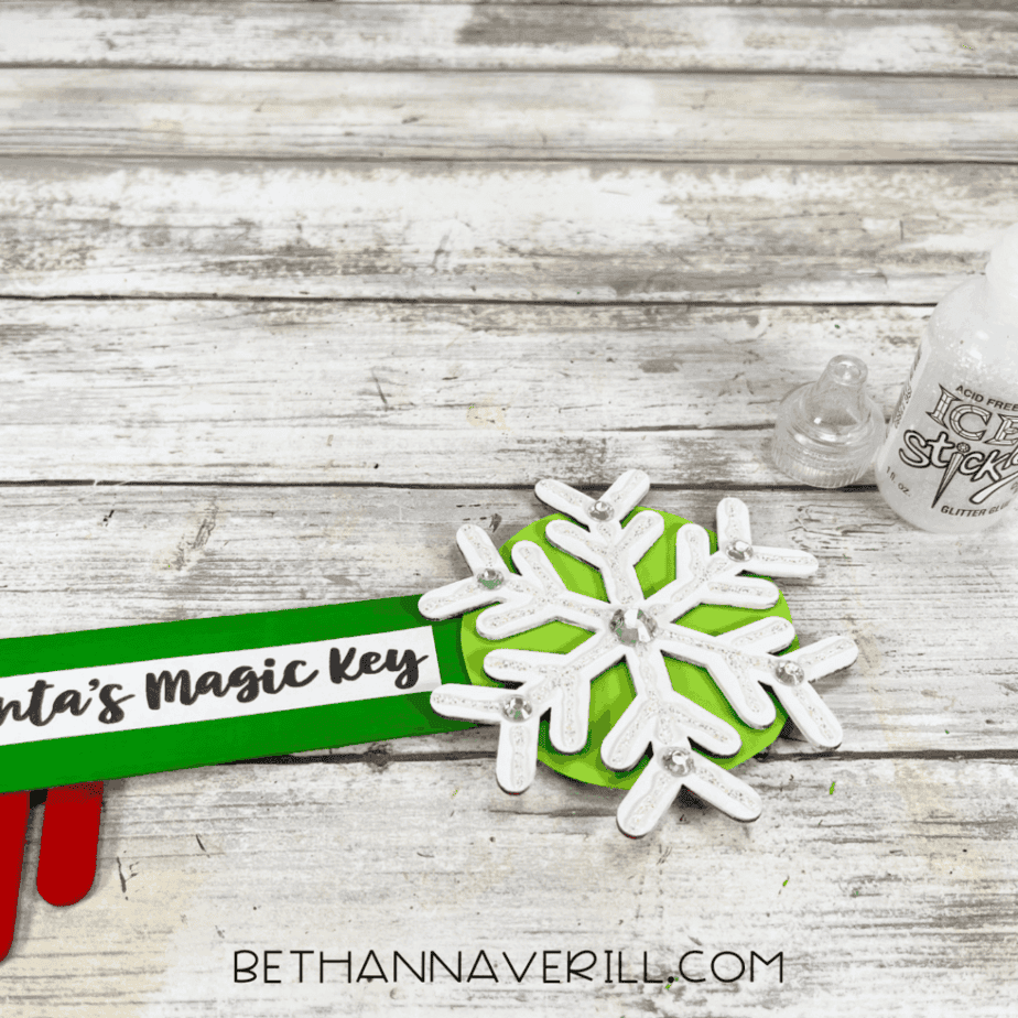 Close-up of a Santa Magic Key craft showing a white snowflake decoration attached to a green popsicle stick key with glitter glue nearby.