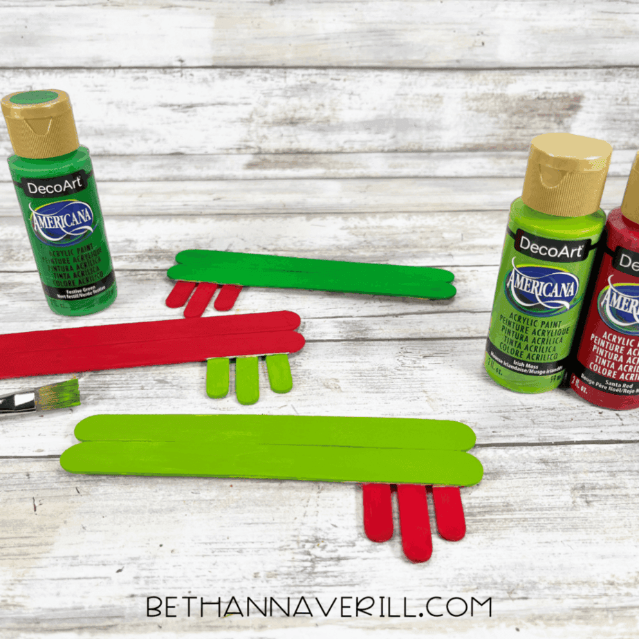 Popsicle stick keys painted red and green drying on a table beside acrylic paint bottles and a paintbrush.