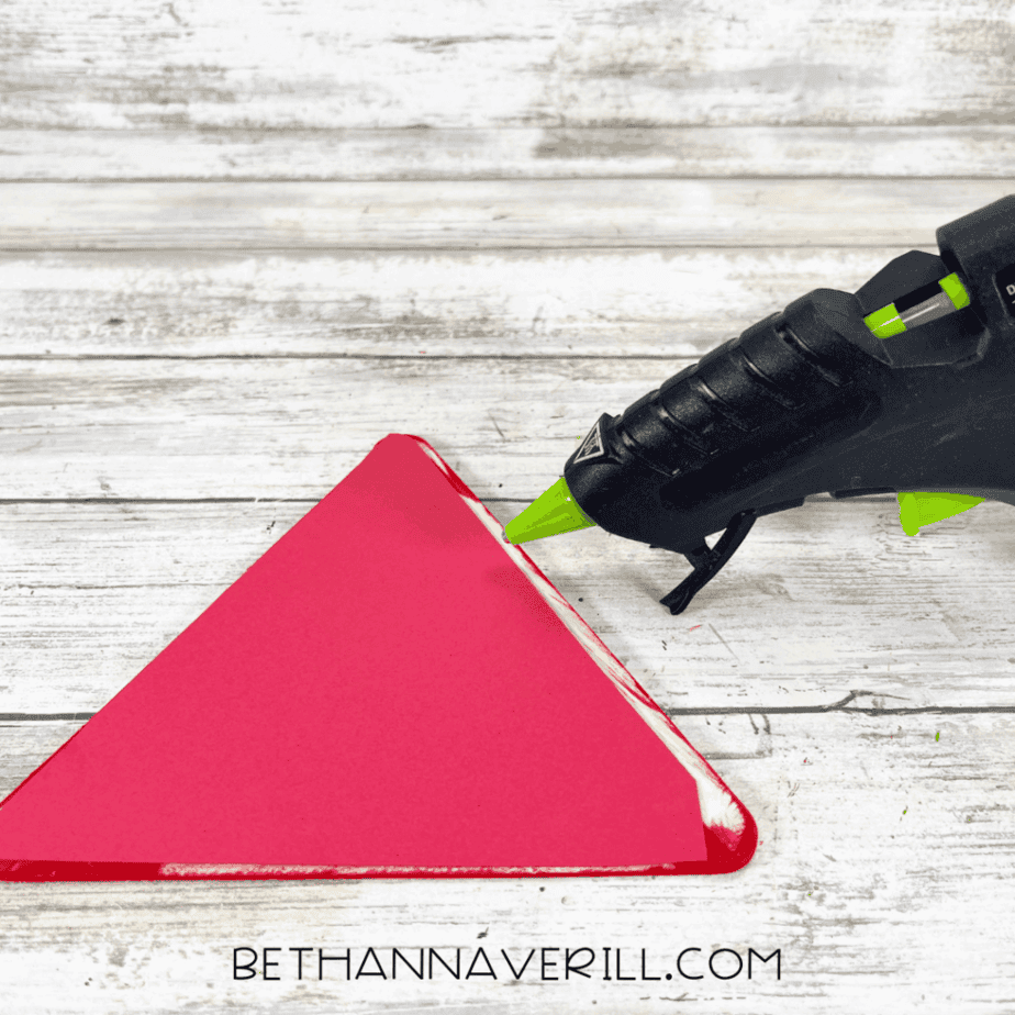 Glue gun applying glue along the edge of a red cardstock triangle on a wood plank background.