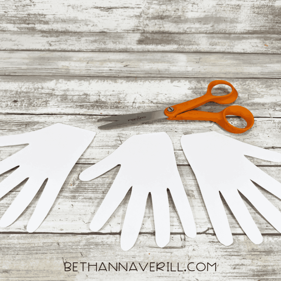 White hand-shaped cutouts arranged on a wood plank surface with scissors above them.