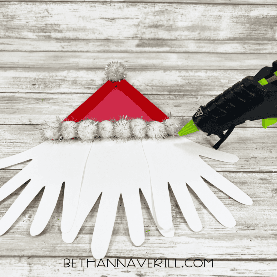 Red craft stick triangle with white pom poms placed above white hand-shaped cutouts while a glue gun applies glue.