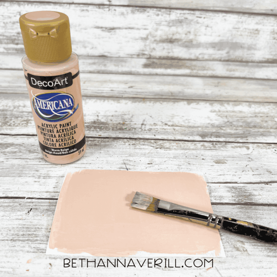 Bottle of beige acrylic paint and paintbrush next to a rectangle of cardstock painted beige.