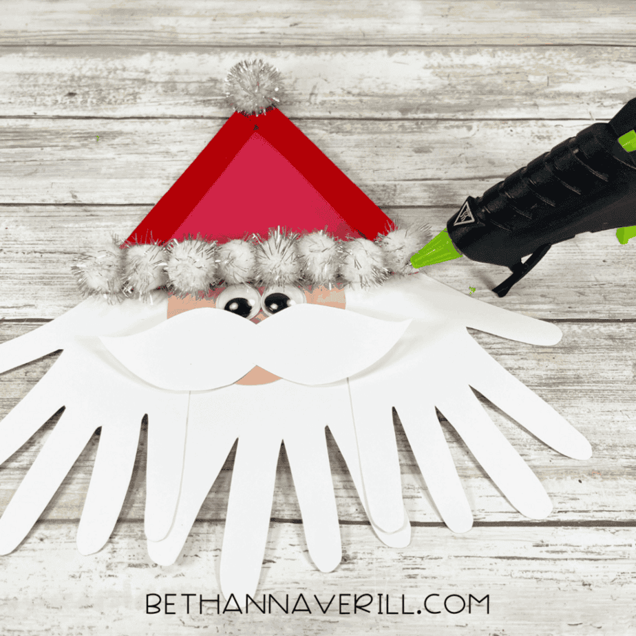 Adding a paper mustache and googly eyes to a paper face beneath a red craft stick triangle while using a glue gun.