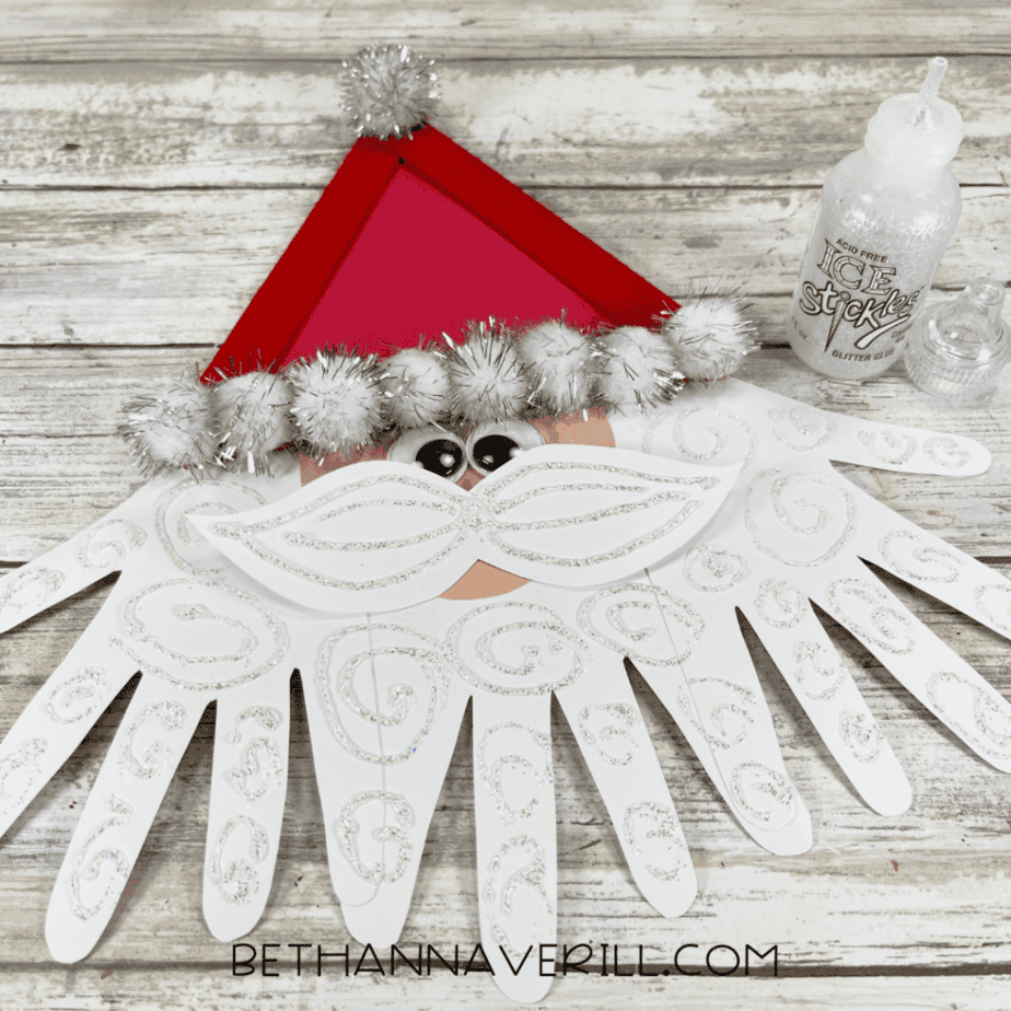 Applying glitter glue to decorate white handprint paper cutouts and a paper mustache on a wood plank surface.