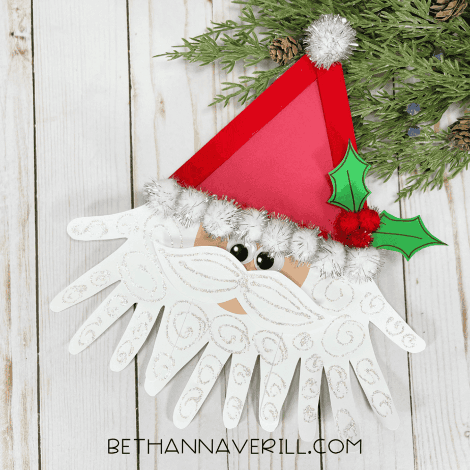 Finished Santa popsicle stick craft with red triangle hat, white pom poms, googly eyes, glitter-decorated paper beard, and green holly leaves on a wood background.