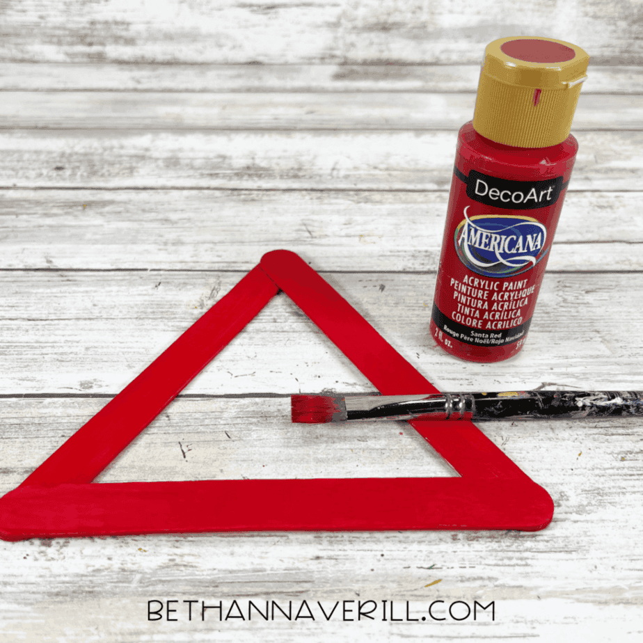 Painting the Santa hat frame red using acrylic paint and a paintbrush on assembled craft sticks.
