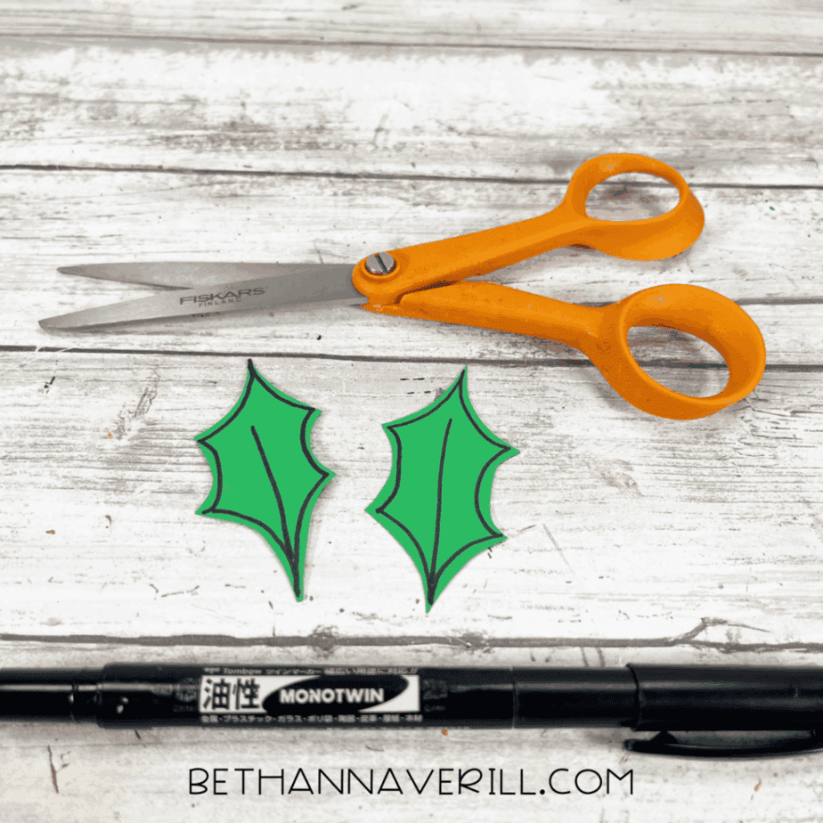 Cut green holly leaves cut from cardstock placed beside scissors and a black marker.