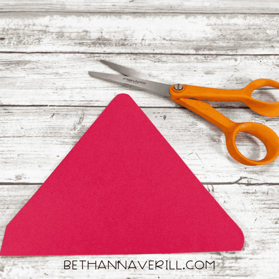 Red cardstock triangle cutout placed on a wood plank surface with orange-handled scissors nearby.