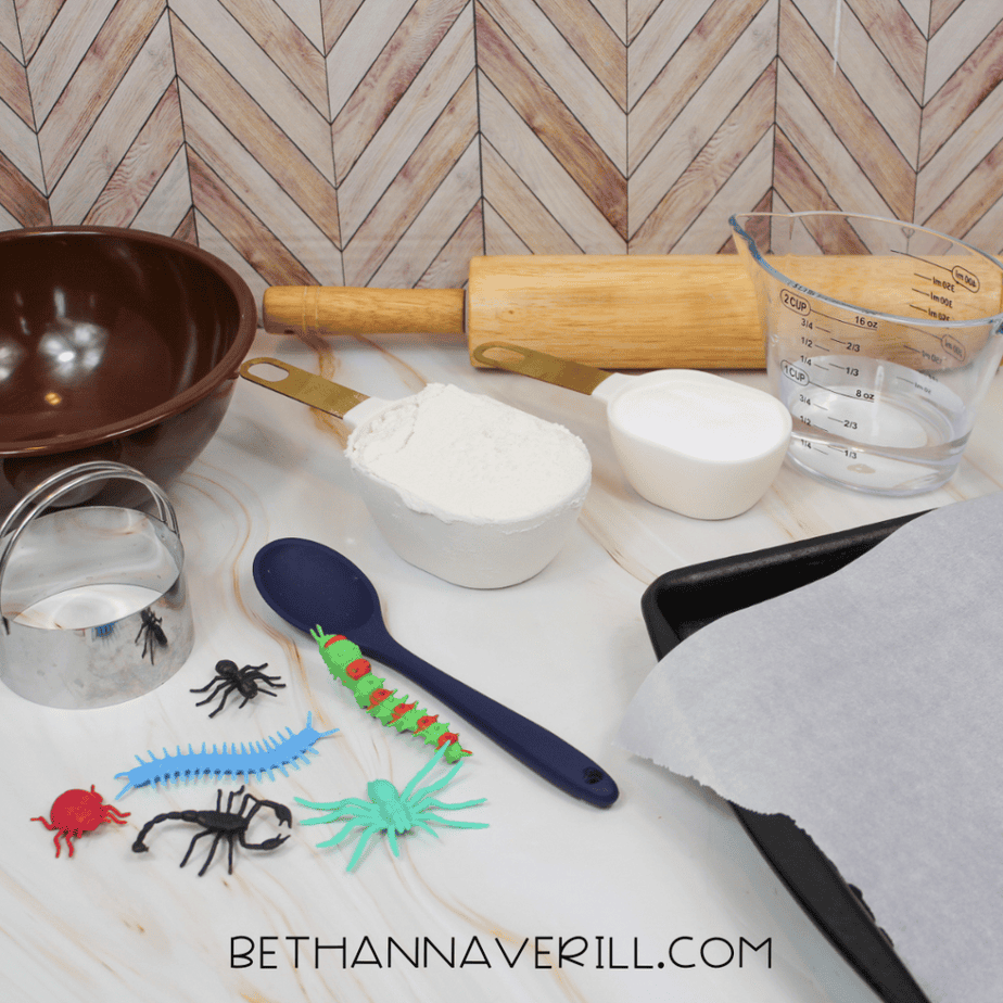 Craft supplies for making salt dough fossils including flour, salt, water, mixing bowl, rolling pin, measuring cups, spoon, parchment paper, baking sheet, and plastic bugs.