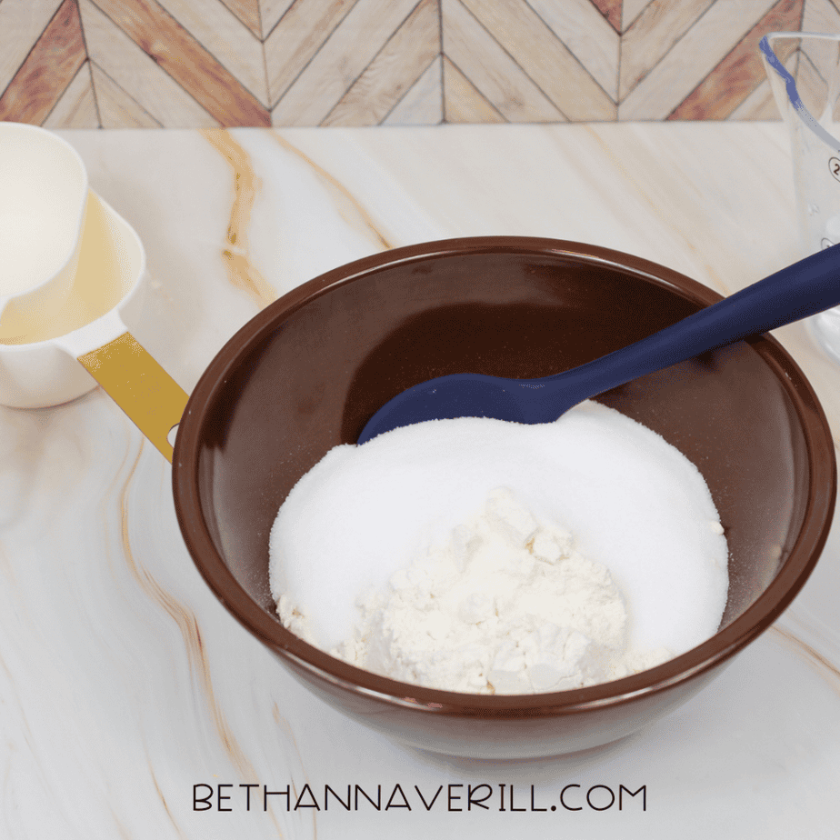 Brown mixing bowl filled with flour and salt with a spoon resting inside, measuring cups nearby on a countertop.