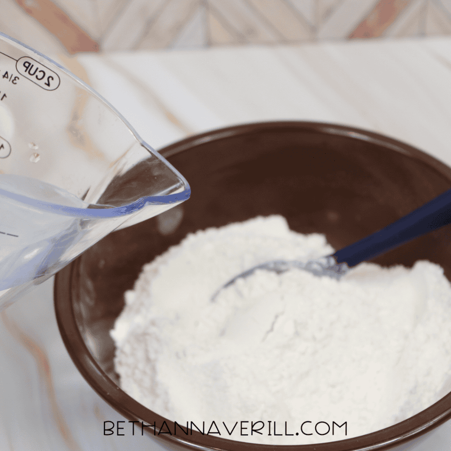 Water being poured from a clear measuring cup into a bowl of flour and salt to make salt dough.