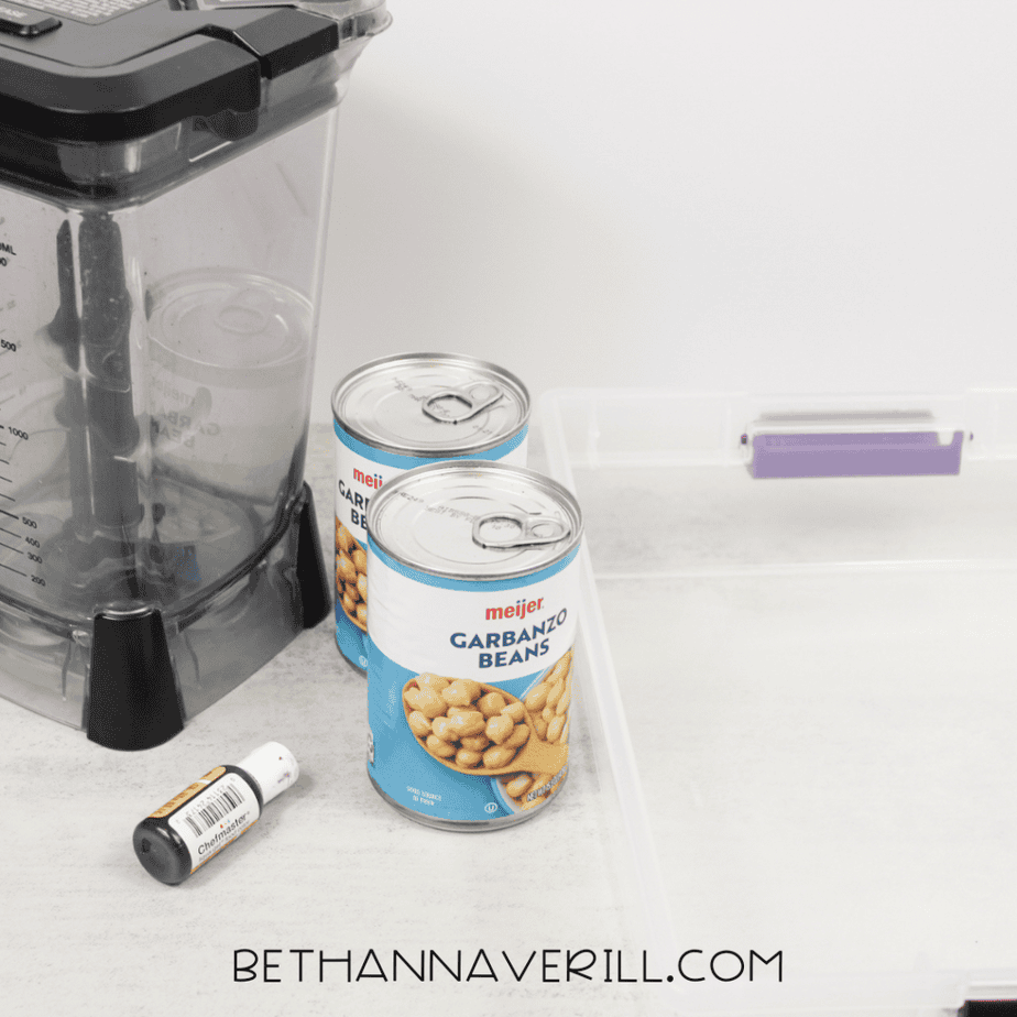 Two cans of garbanzo beans beside a blender, food coloring, and a plastic sensory bin on a countertop for making aquafaba sensory foam.