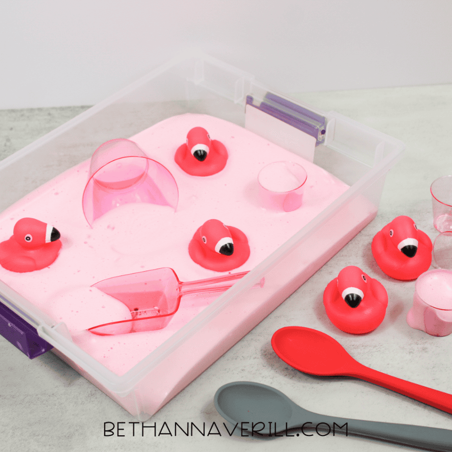 Pink aquafaba foam poured into a sensory bin with small flamingo toys and clear plastic cups.