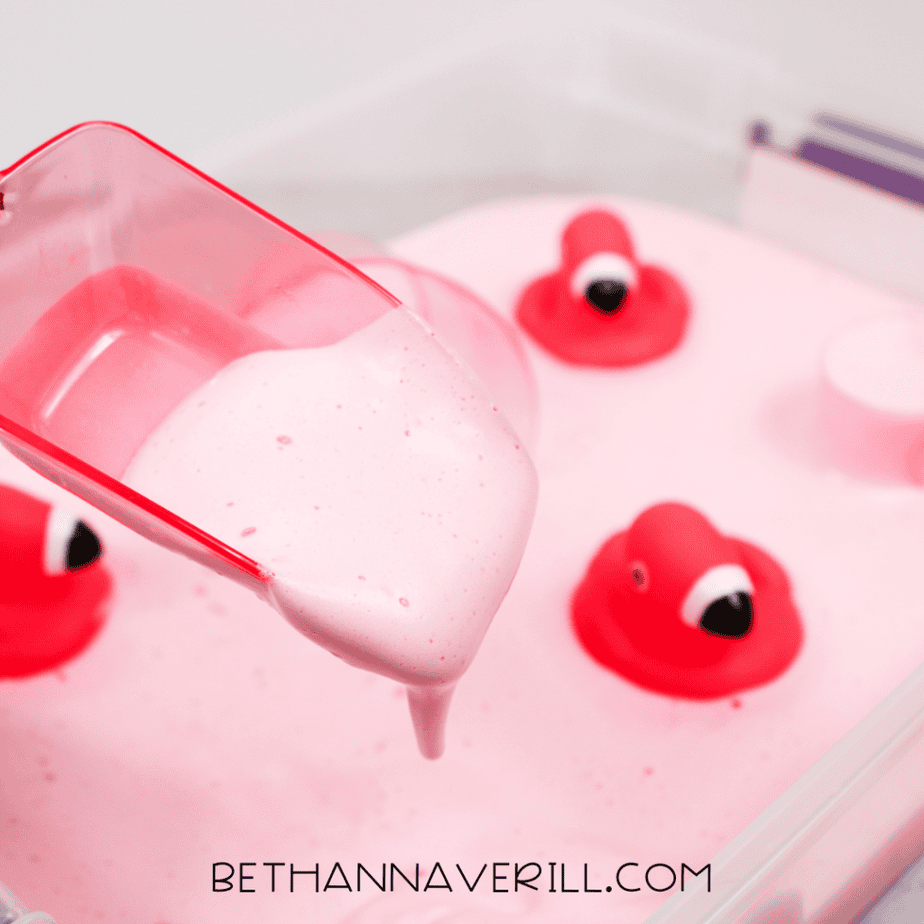 A close-up of a scoop lifting thick, fluffy pink aquafaba foam with flamingo toys in the background.