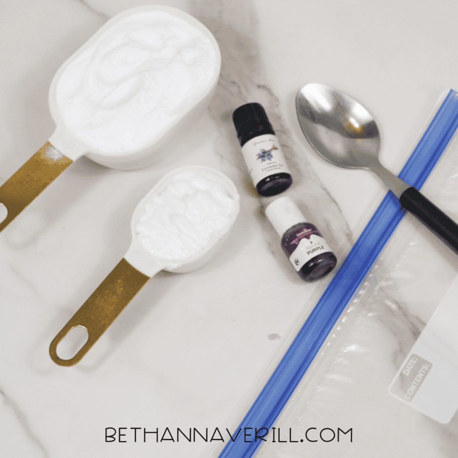 Measuring cups filled with baking soda and lotion alongside lavender essential oil, purple food coloring, a spoon, and a resealable plastic bag on a marble surface.