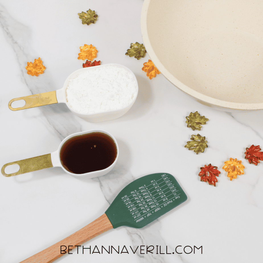 Overhead view of maple syrup slime ingredients including cornstarch in a white measuring cup, maple syrup in a measuring cup, a green spatula, and a large mixing bowl with decorative fall leaf gems on a marble surface.