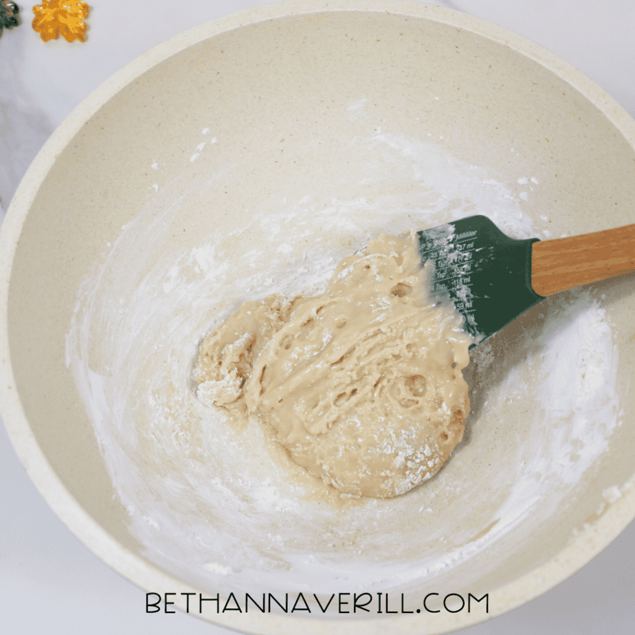 Thickened maple syrup and cornstarch mixture partially combined in a bowl, forming a sticky dough-like consistency as it is stirred with a green spatula.