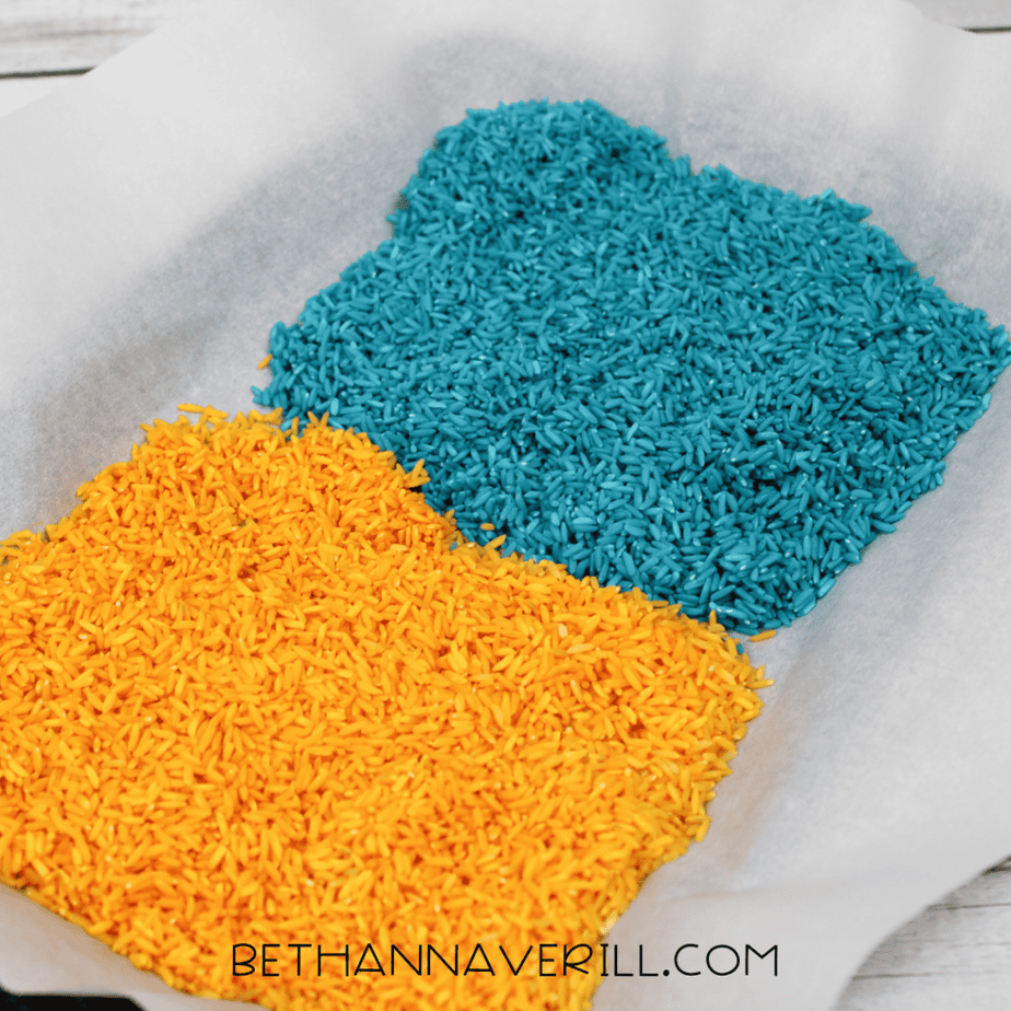 Blue and yellow painted rice spread out on parchment paper to dry on a baking sheet.