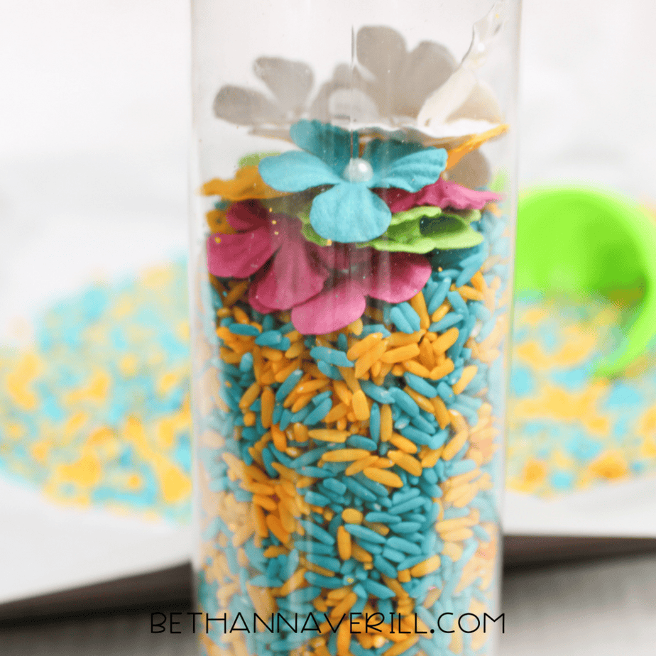 Spring sensory bottle filled with layered blue and yellow rice and colorful faux flowers inside.