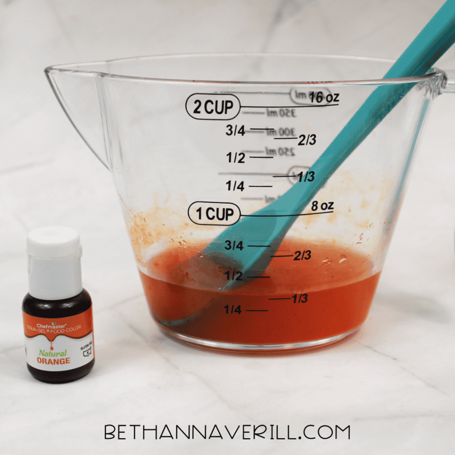 Clear measuring cup filled with orange liquid and a spoon inside, with a small bottle of orange food coloring beside it.
