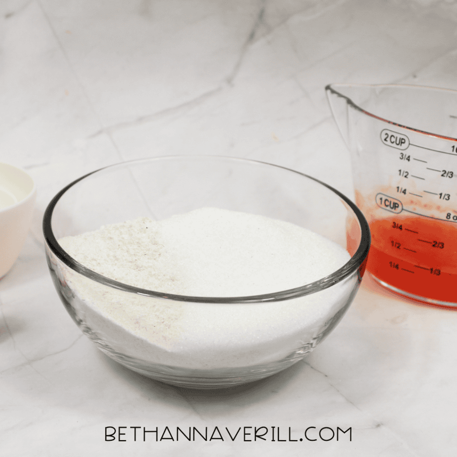 Glass bowl filled with white dry ingredients on a countertop, with a measuring cup and orange liquid mixture nearby.