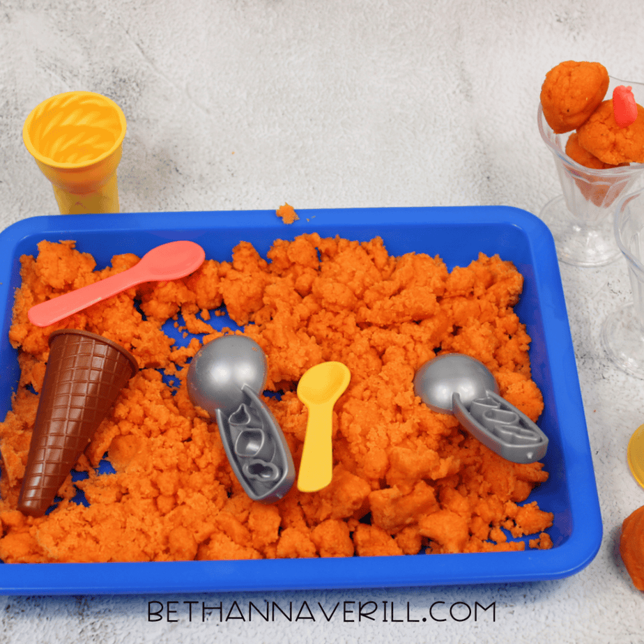 Blue sensory bin filled with bright orange kinetic sand and plastic ice cream scoops and cones.