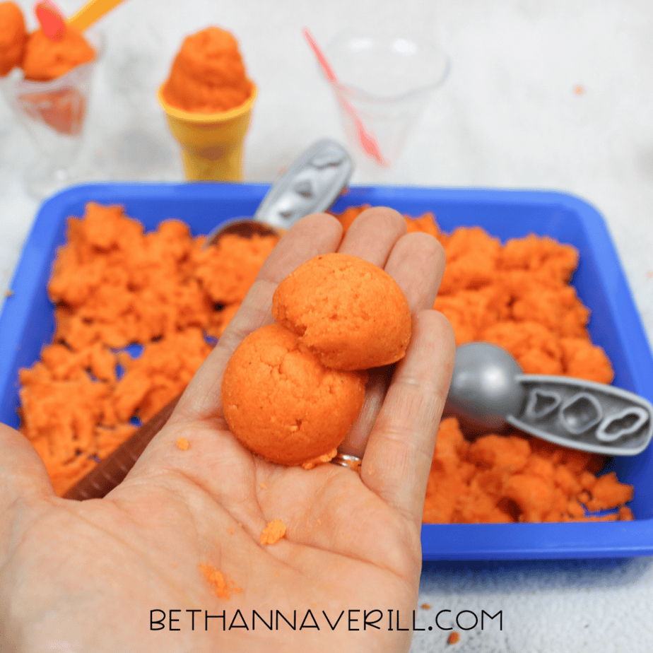 Hand holding two round balls of orange kinetic sand in front of a sensory bin setup.