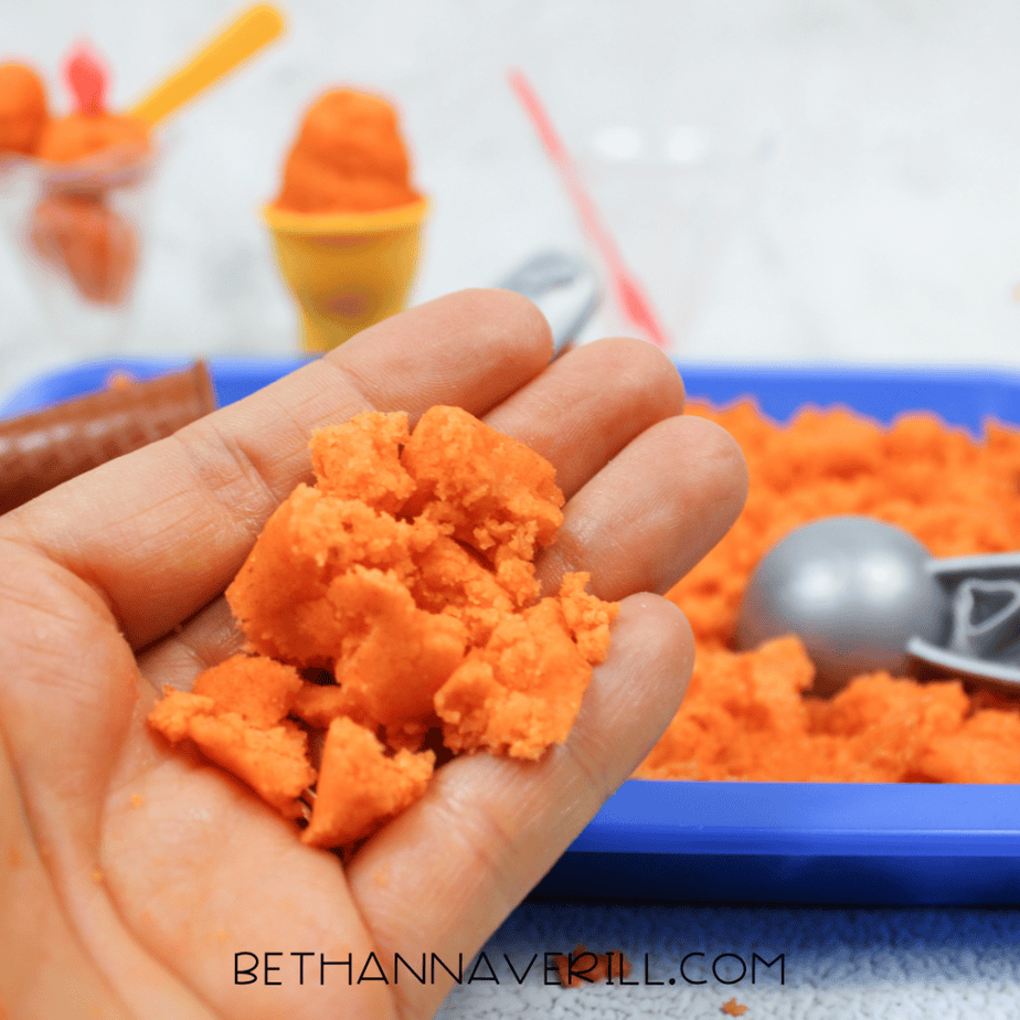 Hand holding a crumbly piece of bright orange kinetic sand above a blue sensory bin.