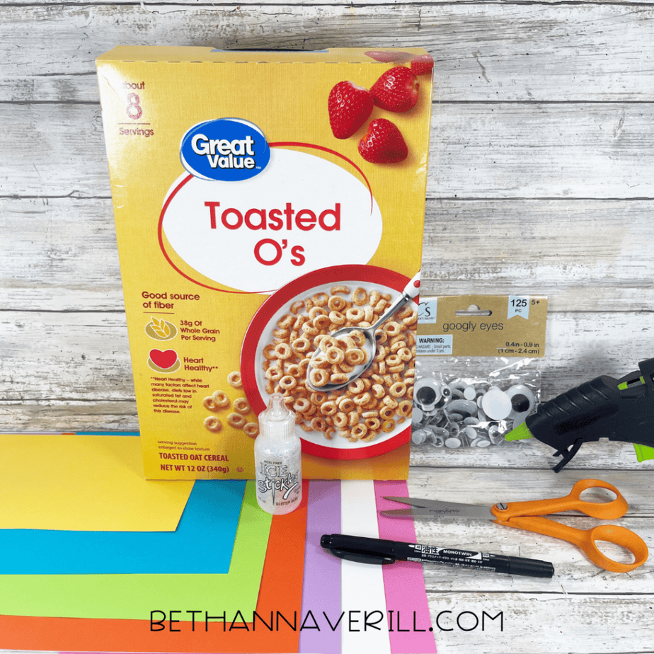 Yellow cereal box labeled “Toasted O’s” displayed with colorful cardstock paper, googly eyes, glue, scissors, and a hot glue gun on a wooden background.