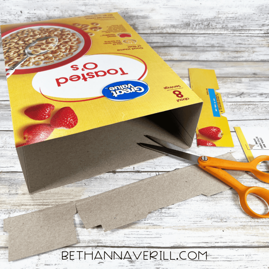 Cereal box laid on its side while scissors cut a long strip from the bottom flap.