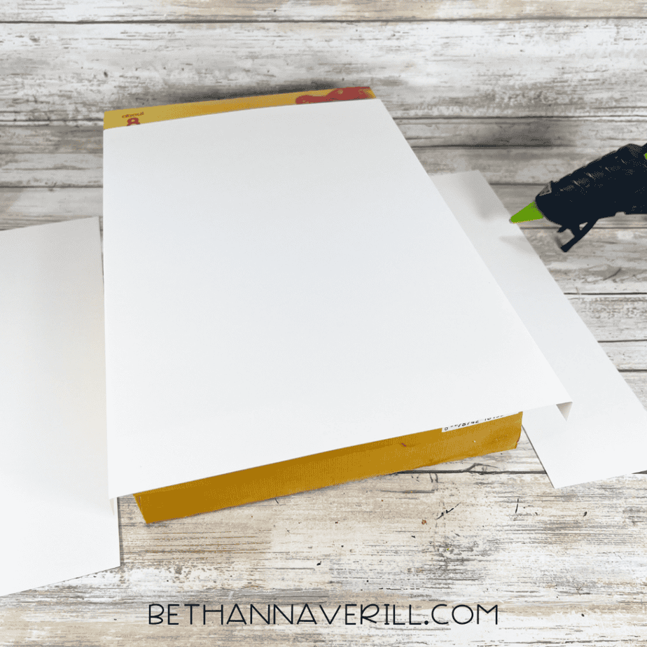 White cardstock being wrapped and glued around the cereal box to cover the printed design.