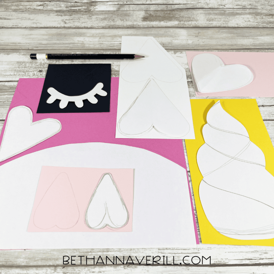 Paper templates of hearts, ears, eyelashes, and a unicorn horn traced onto colored cardstock with scissors nearby.