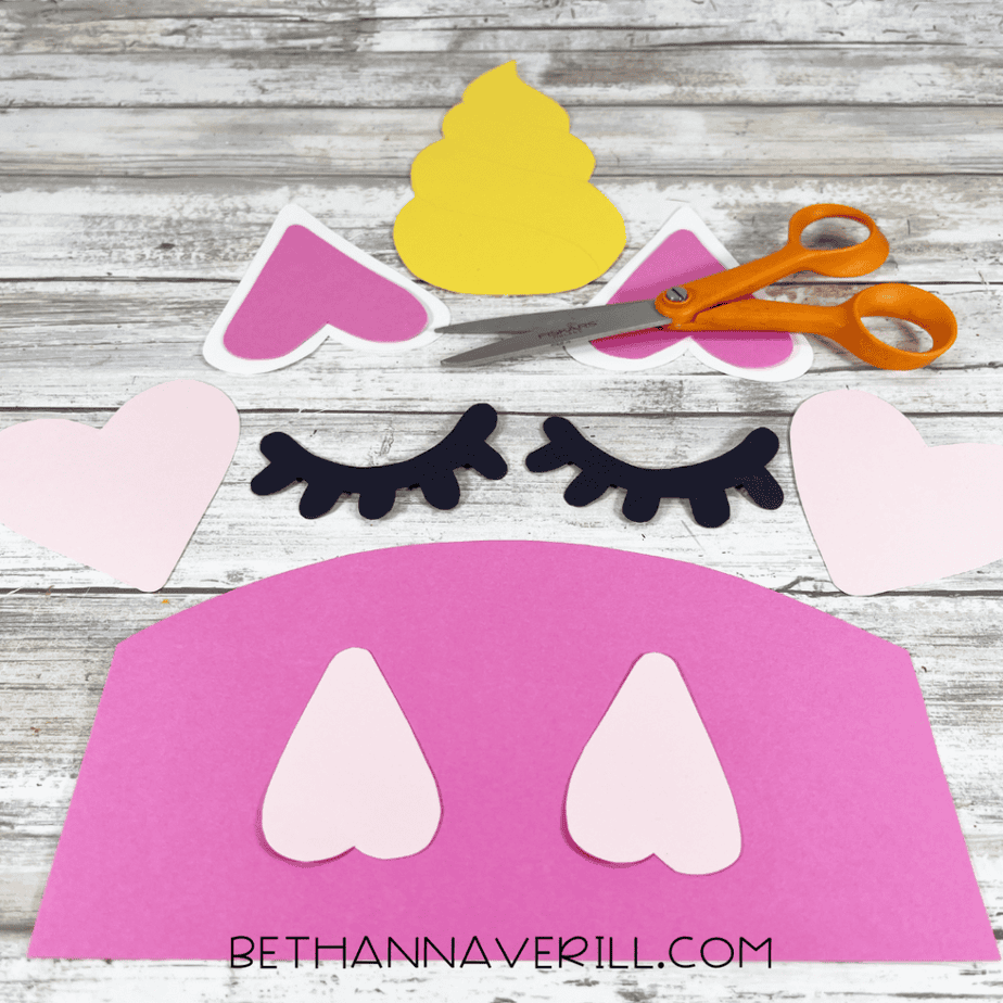Cut out paper pieces including pink ears, yellow horn, black eyelashes, and heart shapes arranged on a table.
