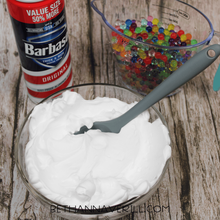 A can of shaving cream next to a glass bowl filled with white foam and a gray spoon. A measuring cup of multicolored water beads sits in the background.