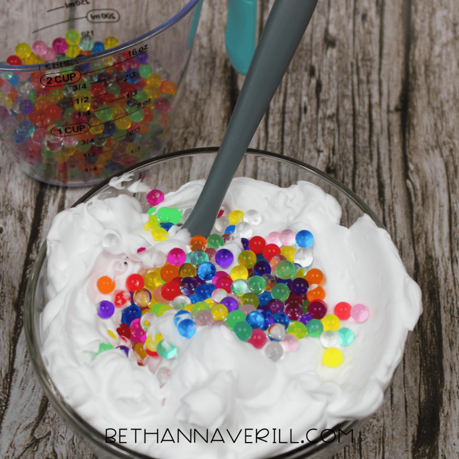 Colorful round water beads poured on top of white foam in a glass bowl with a spoon standing upright in the mixture.